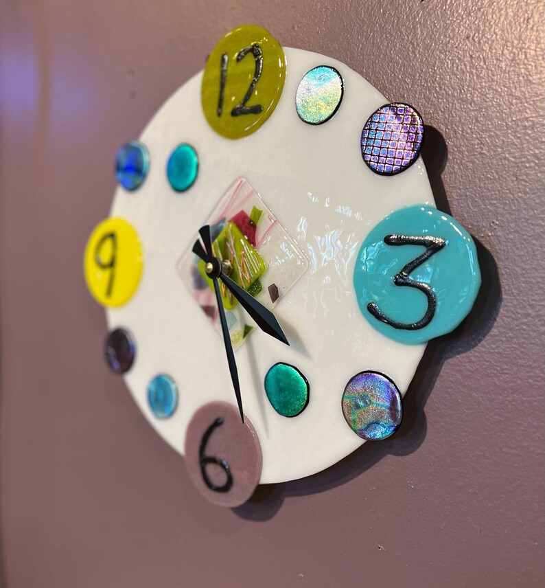 May include: A white clock face with colorful glass accents and black hands. The numbers 12, 9, 3, and 6 are visible. The clock face is surrounded by a white border.