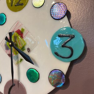 May include: A white clock face with colorful glass accents. The clock face has a black hour hand and minute hand. The number three is written in black on a turquoise circle.