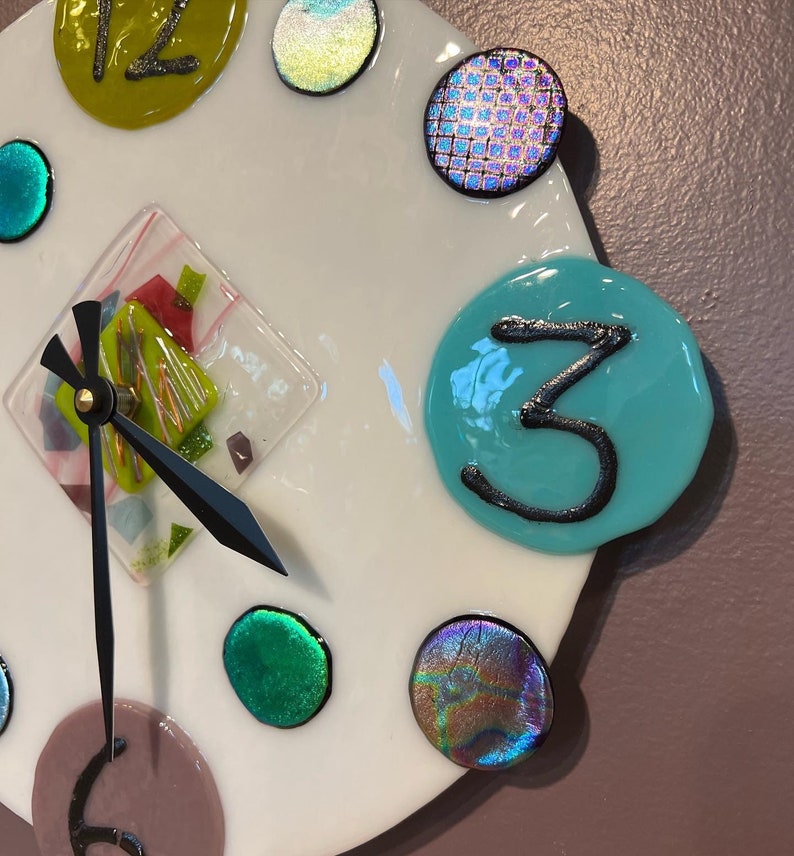 May include: A white clock face with colorful glass accents. The clock face has a black hour hand and minute hand. The number three is written in black on a turquoise circle.