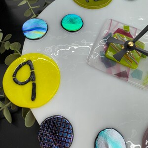 May include: A white clock face with yellow, green, blue, and purple glass accents. The numbers 9 and 12 are visible on the clock face.