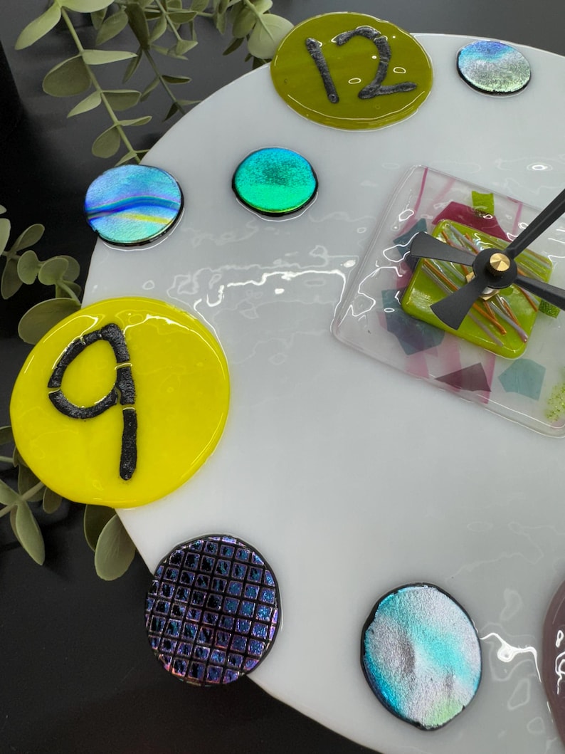 May include: A white clock face with yellow, green, blue, and purple glass accents. The numbers 9 and 12 are visible on the clock face.