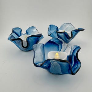 May include: Three glass candle holders with a wavy, ruffled edge design. Each holder features a marbled pattern of deep blue, light blue, and white swirls. Inside each holder sits a small white tea light with a glowing yellow LED flame.