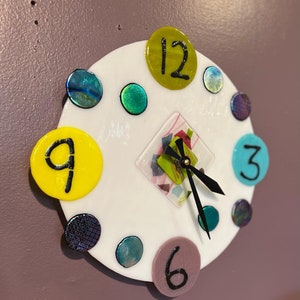 May include: A white circular clock with black hands and colorful glass accents. The numbers 12, 9, 6, and 3 are visible on the clock face.