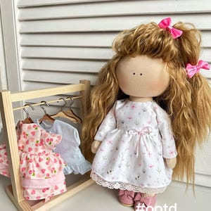 May include: A doll with long curly brown hair wearing a white dress with pink flowers and a pink bow in her hair. The doll is standing next to a wooden doll clothes rack with two pink and white dresses hanging on it. The text #ootd is written on the floor below the doll.