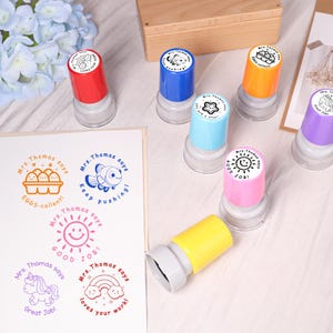 May include: A collection of colorful, self-inking stamps with various designs and phrases. The stamps feature images of a fish, eggs, a sun, a unicorn, and a rainbow, along with the phrases "Good Job!", "Great Job!", and "Loves your work!"