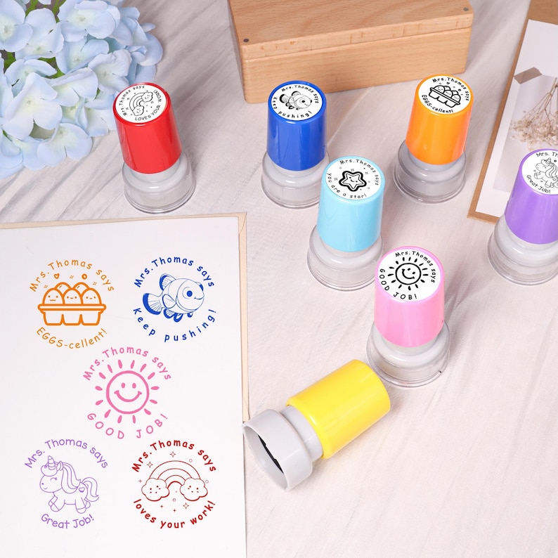 May include: A collection of colorful, self-inking stamps with various designs and phrases. The stamps feature images of a fish, eggs, a sun, a unicorn, and a rainbow, along with the phrases "Good Job!", "Great Job!", and "Loves your work!"