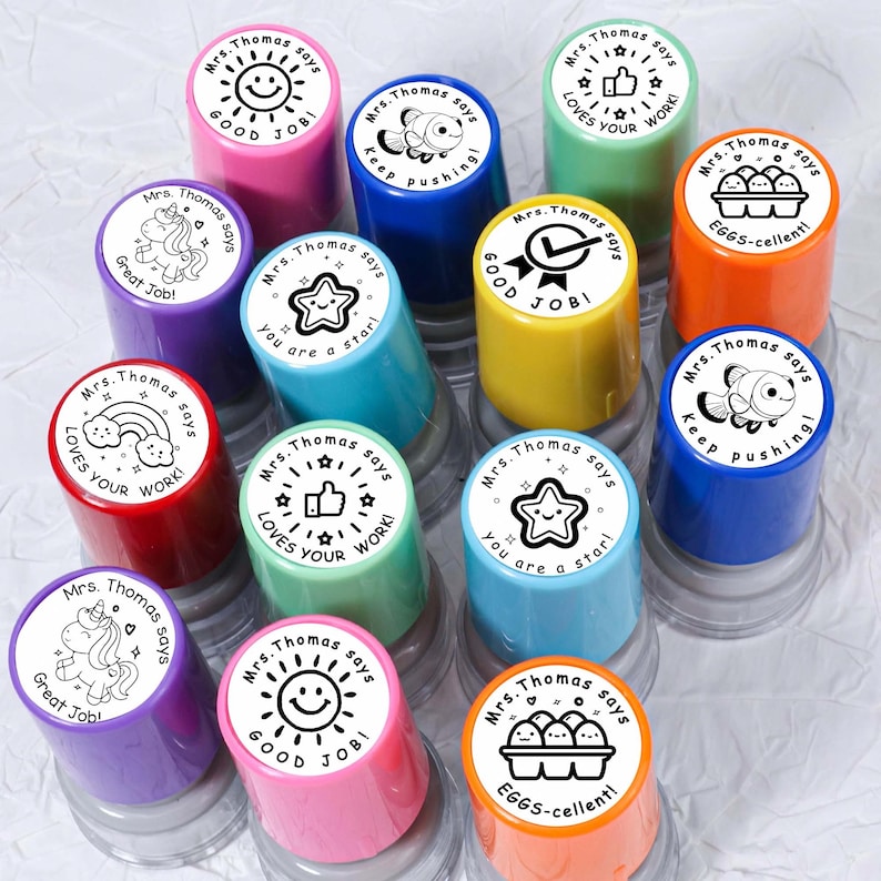 May include: A collection of colorful, round stamps with various designs and phrases. Each stamp has a different color and features a unique image, such as a star, smiley face, or thumbs up, along with the text "Mrs. Thomas says" and a positive message.