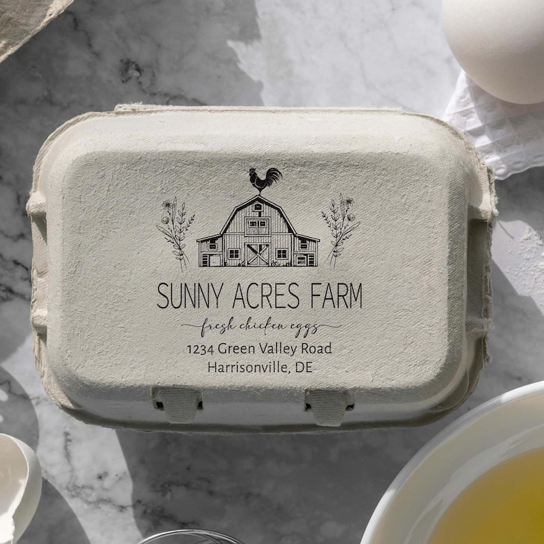 May include: A white egg carton with a black and white illustration of a farmhouse with a rooster on top. The text "Sunny Acres Farm" is printed in black, along with the phrase "fresh chicken eggs" in cursive. The address "1234 Green Valley Road, Harrisonville, DE" is also printed in black.