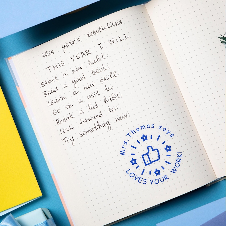 May include: Open notebook with handwritten resolutions, including "Start a new habit" and "Read a good book." A blue stamp reads "Mrs. Thomas says LOVES YOUR WORK!" on the page. The notebook is on a blue surface.