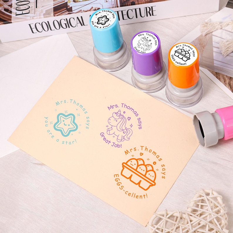 May include: Three colorful stampers with the text "Mrs. Thomas says" and various designs, including a star, a unicorn, and eggs. The stamps are blue, purple, and orange. The stamped images on the paper say "You are a star!", "Great Job!", and "EGGS-cellent!"