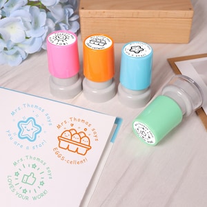 May include: A collection of colorful stamps with the text "Good Job!", "Eggs-cellent!", and "You are a star!" on the stamp heads. The stamps are pink, orange, blue, and green. A sheet of paper with matching stamp designs is also visible.