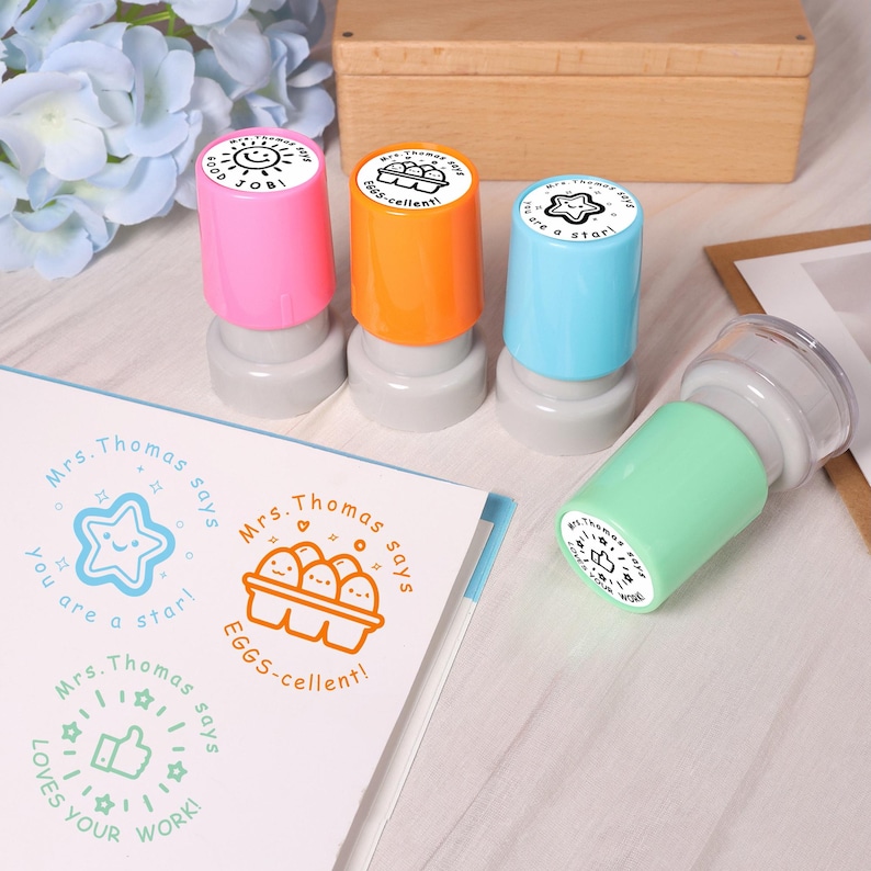 May include: A collection of colorful stamps with the text "Good Job!", "Eggs-cellent!", and "You are a star!" on the stamp heads. The stamps are pink, orange, blue, and green. A sheet of paper with matching stamp designs is also visible.