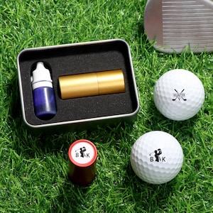 May include: A golf set on green grass. The set includes two white golf balls with black logos, a gold-coloured cylindrical item, a small bottle of blue liquid, and a red and white stamp. A golf club is also visible.