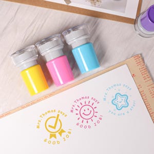 May include: Three colorful stampers with clear tops and gray handles. The stampers are yellow, pink, and blue. The stamped images say "Mrs. Thomas says" and "Good Job!" with a checkmark, sun, and star.