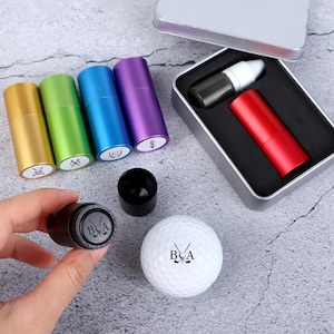 May include: A golf ball and marker set. The set includes a white golf ball with the letters "BCA" and crossed golf clubs, a black marker, and several coloured markers in gold, green, blue, and purple.