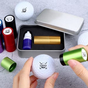 May include: Golf ball marking kit with various coloured stampers, ink, and a golf ball. The kit includes a silver metal case and a gold-coloured stamper. The golf balls have custom logos.