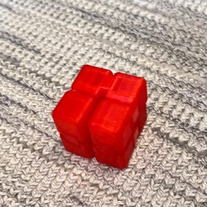 May include: A vibrant red, three-dimensional cube-shaped object with a textured surface. The object is placed on a grey, knitted fabric background. The cube appears to be made of plastic.