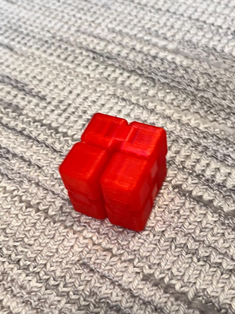 May include: A vibrant red, three-dimensional cube-shaped object with a textured surface. The object is placed on a grey, knitted fabric background. The cube appears to be made of plastic.