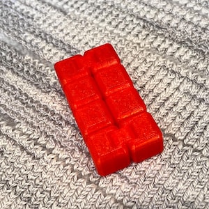 May include: A vibrant red wax melt, rectangular in shape with a block design. It is positioned on a grey, knitted fabric background. The wax melt is a solid, bright colour.