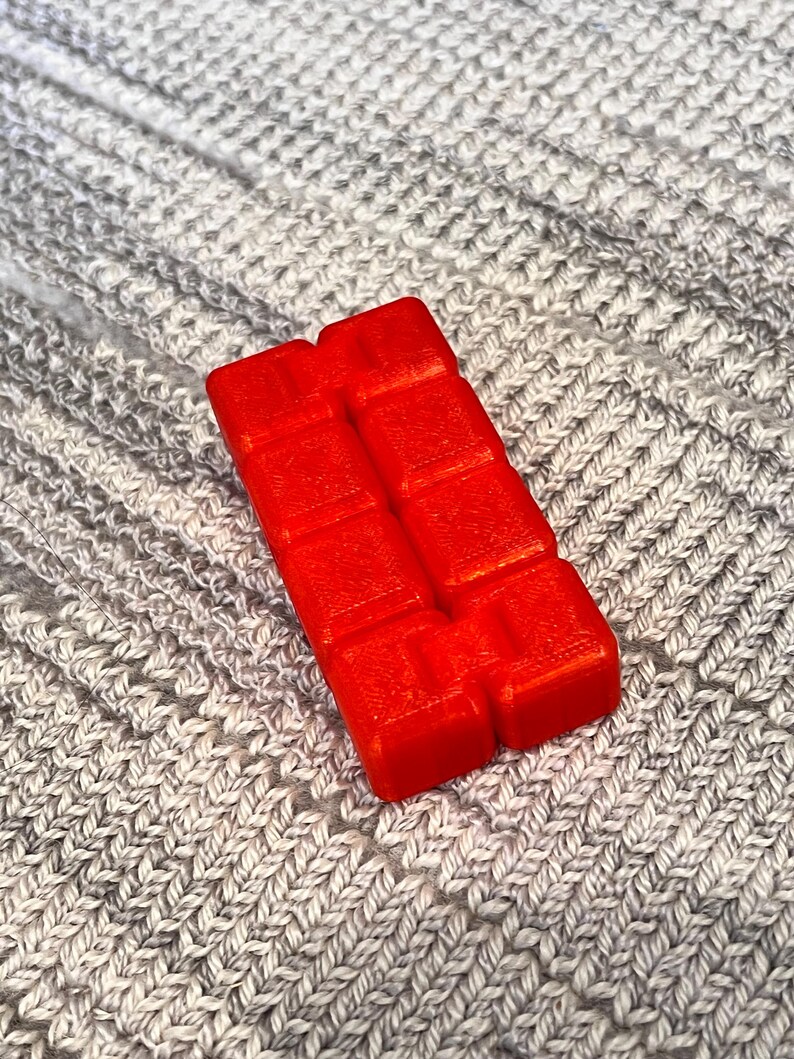 May include: A vibrant red wax melt, rectangular in shape with a block design. It is positioned on a grey, knitted fabric background. The wax melt is a solid, bright colour.