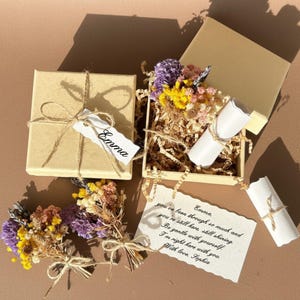 May include: A square cardboard gift box with a twine bow and a tag labelled Emma, alongside an open box containing dried purple and yellow flowers, a white paper scroll, and a note with the text: Emma, you've been through so much and you're still here, still shining. Be gentle with yourself. I'm right here with you. With love, Sophia.
