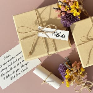 Könnte beinhalten: Zwei in braunes Papier verpackte Geschenke, mit Schnur gebunden, eines mit einem Etikett "Olivia". Ein kleiner Strauß getrockneter Blumen in Gelb, Lila und Rosa liegt daneben. Eine handschriftliche Notiz lautet "Olivia, is new chapter, remem..."