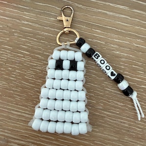 May include: A ghost-shaped keychain crafted from white pony beads with two black beads for eyes. A gold-tone lobster clasp is attached to the top. A dangling strand features black and white letter beads spelling BOO with a heart symbol.