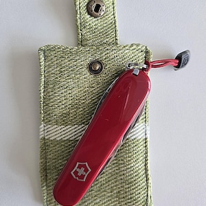 May include: A red Swiss Army knife with a silver cross emblem, resting in a green fabric case with a snap closure. The case has a woven texture with white stripes. A red cord with a black end is attached to the knife.