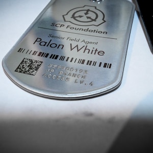 K&ouml;nnte beinhalten: Ein silbernes Metallschild mit dem Text "SCP Foundation" und "Senior Field Agent Palon White" darauf gedruckt. Das Schild hat auch einen Barcode und den Text "#26300193 JIP BRANCH ACCESS LV-4".