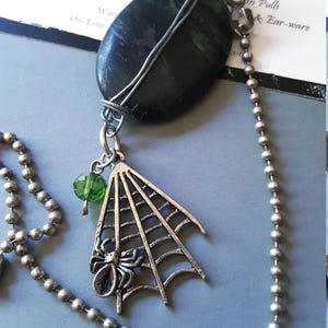 Puede incluir: Un collar de cadena de bolas plateada con una piedra ovalada de color verde oscuro envuelta en alambre. Un dije plateado de una araña en su telaraña cuelga de la piedra, junto a una pequeña cuenta verde translúcida facetada. Tarjeta: Wanna-B-RockStar.