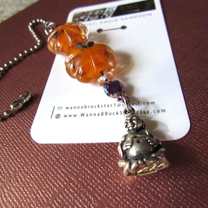 May include: A decorative chain with two orange pumpkin beads, a purple and clear bead, and a silver Buddha charm. The chain is attached to a white tag with the text "JUST ANGIE SAMPSON" and contact information.