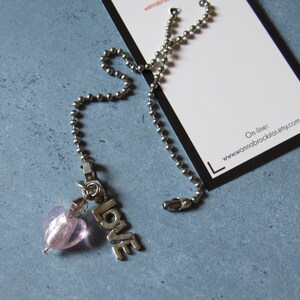 May include: A silver-toned chain with a pink heart-shaped bead and the word "LOVE" in silver letters. A white tag with black border is in the background. The tag has the website address "www.annabrockstar.etsy.com".