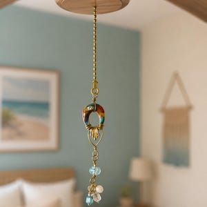 Könnte beinhalten: Eine Deckenventilator-Zugkettenverlängerung mit einer Messing-Kugelkette, einem gebogenen, mehrfarbigen, hufeisenförmigen Keramik-Anhänger mit Golddrahtwicklung und einem Bündel aus hellblauen, klaren und weißen Perlen am unteren Ende.