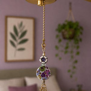 May include: A gold-toned metal ceiling fan pull chain featuring a small white and purple flower-shaped bead, a larger circular flat bead with a multi-colored floral pattern in shades of purple, green, and blue, and three small dangling crystal beads.