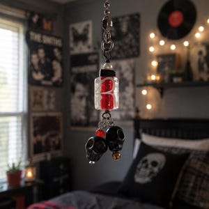 Può includere: Un'estensione per catenella di ventilatore a soffitto con una perla di vetro rettangolare trasparente contenente un interno rosso e una lettera R bianca. L'ornamento include due piccole perle a forma di teschio nero, una rossa e una dorata.