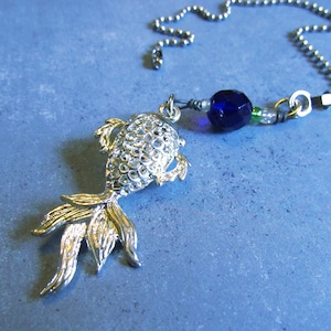 Puede incluir: Colgante de pez plateado con aletas y cola doradas, sujeto a una cadena plateada. Una cuenta facetada azul oscuro y cuentas verdes y transparentes más pequeñas también están unidas. Fondo azul texturizado.