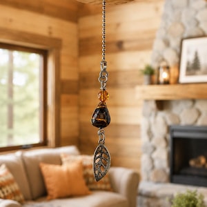 May include: A decorative hanging ornament featuring a silver-toned ball chain attached to a wooden base. The ornament includes a faceted amber-colored bead, a dark brown teardrop-shaped glass bead with swirl patterns, and a silver-toned leaf charm.