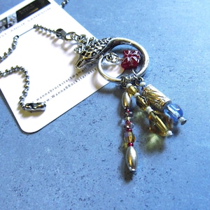 May include: A silver-coloured keychain featuring a dragon head, a red flower, and dangling beads in gold, red, and blue. The keychain is attached to a ball chain and a white tag with text.