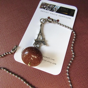 Puede incluir: Un collar con un colgante de la Torre Eiffel plateado y una gran cuenta redonda de piedra marrón. El collar está sujeto a una cadena de bolas. El nombre del artista, Angie Sampson, está impreso en la etiqueta.
