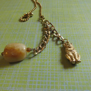 May include: A gold-toned necklace featuring a chain with a large, light-colored stone pendant and a small, detailed cat charm. The cat charm is in a sitting position. The necklace is displayed on a green patterned surface.