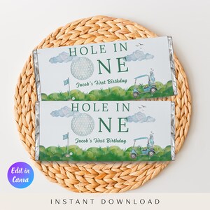 Pode incluir: Duas embalagens de barras de chocolate com tema de golfe. As embalagens apresentam o texto "HOLE IN ONE" e "Jacob's First Birthday". O design inclui uma bola de golfe, um carrinho de golfe e relva verde. As embalagens estão sobre um sousplat circular tecido.