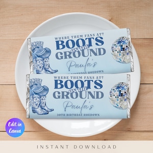May include: Two light blue candy bar wrappers with silver foil edges, featuring a cowboy boot and hat illustration, a disco ball, and the text "BOOTS ON THE GROUND." The wrappers are on a white plate.