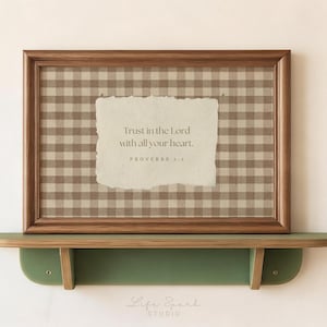May include: A rectangular wooden frame containing a beige and brown gingham patterned background with a centered, torn-edge paper insert featuring the text: Trust in the Lord with all your heart. PROVERBS 3:5. The frame rests on a green wall-mounted shelf.