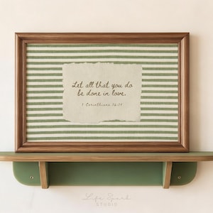 Può includere: Una cornice rettangolare in legno contenente un inserto in tessuto con strisce orizzontali verde salvia e bianche. Al centro c'è un ritaglio di carta color crema con il testo: Let all that you do be done in love. 1 Corinthians 16:14. Poggia su una mensola verde.