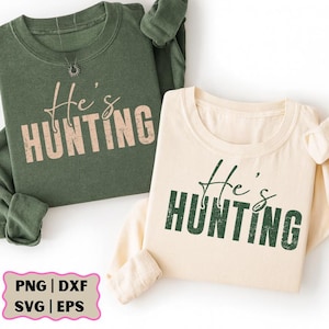 K&ouml;nnte beinhalten: Zwei lang&auml;rmlige Sweatshirts, eines olivgr&uuml;n und eines cremefarben, mit dem Aufdruck "He's HUNTING" auf der Vorderseite. Der Text ist in einer Used-Schriftart gehalten. Die Sweatshirts sind wahrscheinlich f&uuml;r ein Paar oder eine Familie.