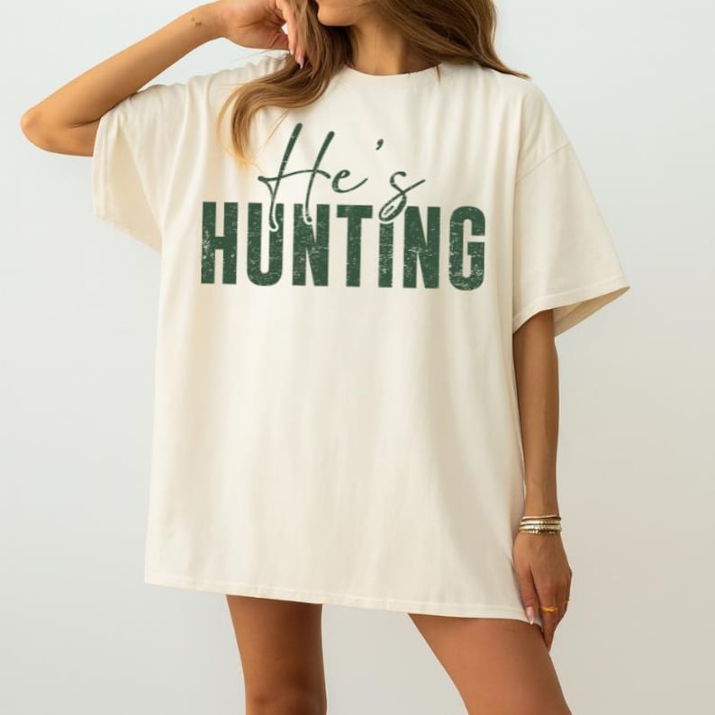 K&ouml;nnte beinhalten: Cremefarbenes T-Shirt mit dem Text "He's Hunting" in gr&uuml;nem, verwaschenem Schriftzug. Das Shirt hat kurze &Auml;rmel und eine lockere Passform, geeignet f&uuml;r Freizeitkleidung.