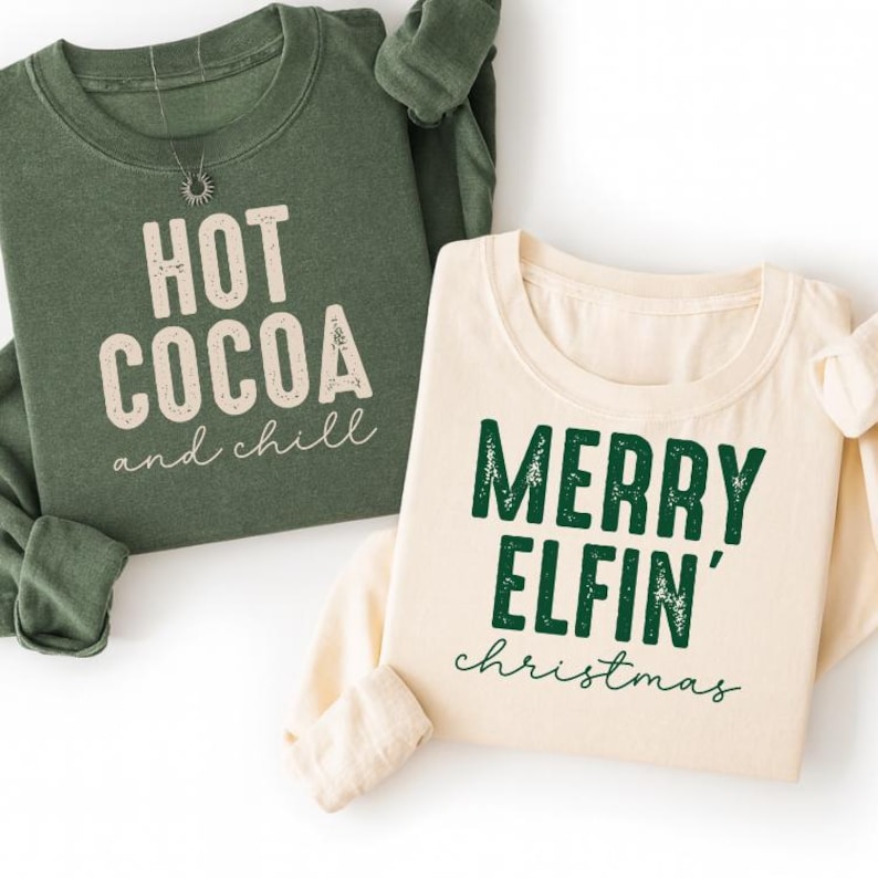 May include: Two long-sleeved sweatshirts. One is olive green with the text "HOT COCOA and chill" in white. The other is cream-colored with the text "MERRY ELFIN' christmas" in green. Both are casual and festive.