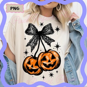 May include: Cream-colored t-shirt featuring a black bow and two orange jack-o'-lanterns with smiling faces. The design includes black star accents, perfect for Halloween. The shirt has a relaxed fit.