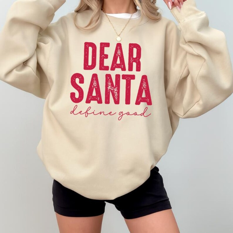 May include: A beige sweatshirt with the text "DEAR SANTA define good" in red. The sweatshirt is a neutral color and has long sleeves. The text is in a bold, distressed font.
