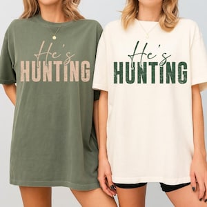 K&ouml;nnte beinhalten: Zwei kurz&auml;rmelige T-Shirts. Eines ist olivgr&uuml;n mit dem Text "He's HUNTING" in Hellbraun. Das andere ist cremefarben mit dem gleichen Text in Dunkelgr&uuml;n. Beide Shirts haben einen Rundhalsausschnitt.